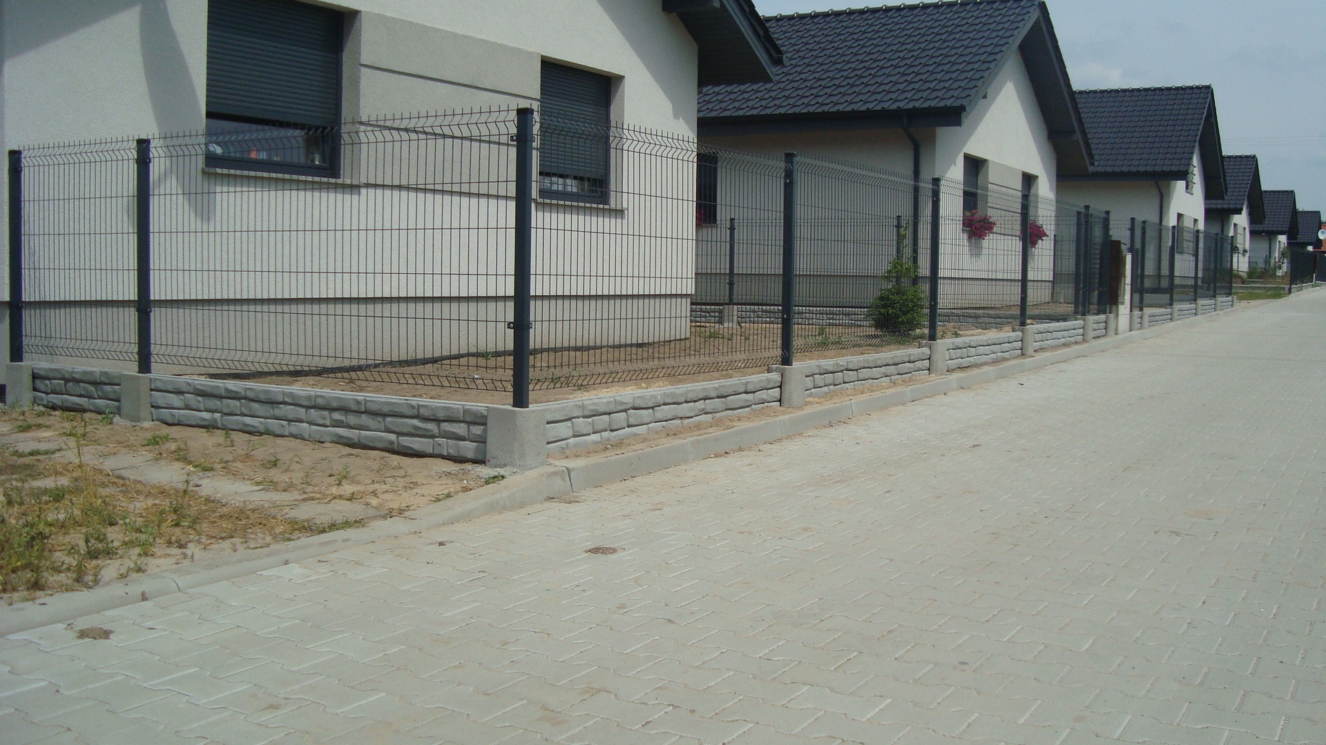 Długa linia ogrodzeń panelowych z podmurówką z szarej cegły otacza szereg identycznych domów z ciemnymi dachami i zamkniętymi roletami, widziana z perspektywy chodnika z kostki brukowej.