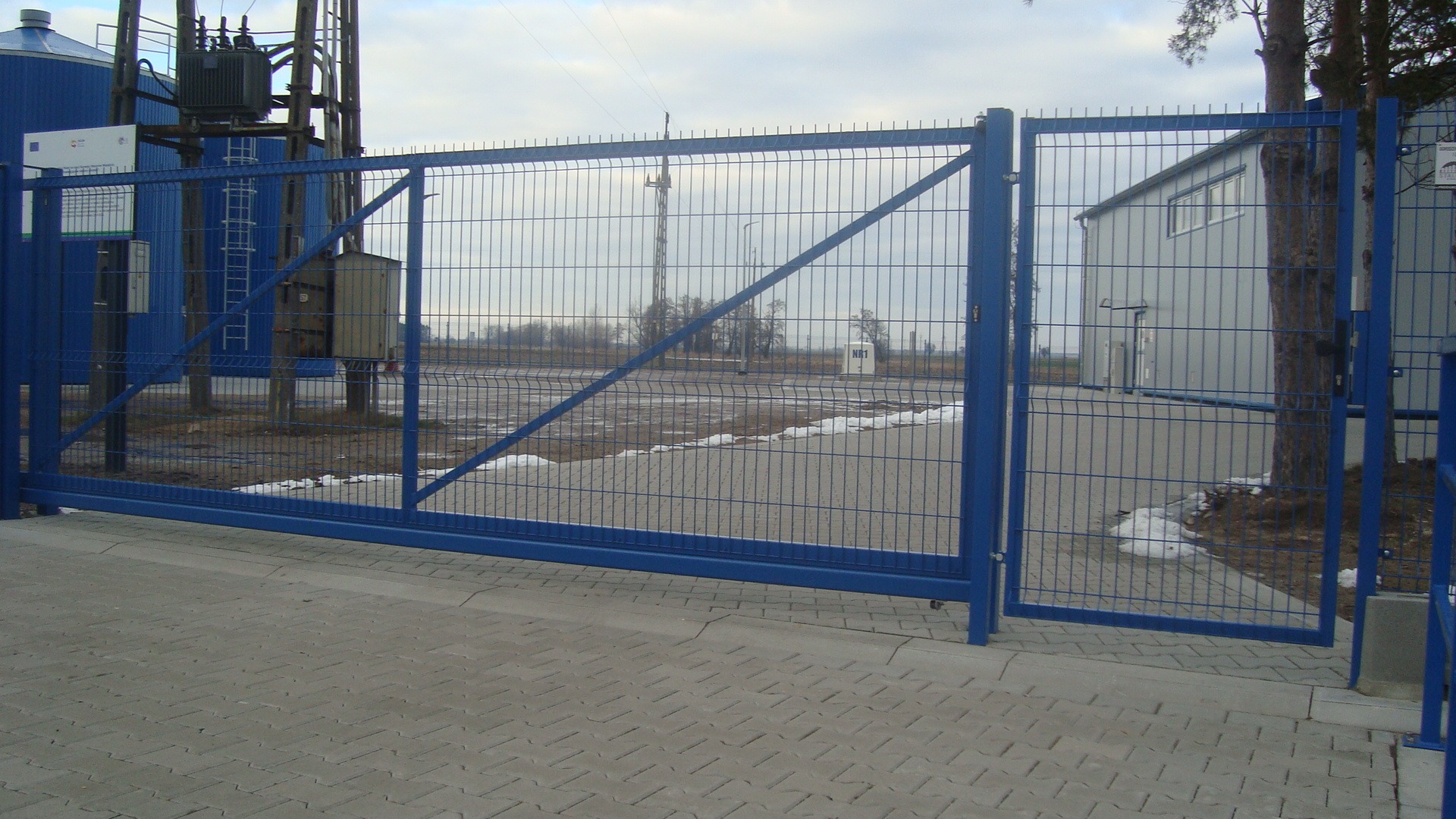 Przesuwna brama wjazdowa z paneli zgrzewanych, pomalowana na niebiesko, widok z zewnątrz na teren przemysłowy i budynek gospodarczy w tle, fragmenty topniejącego śniegu przy bramie.