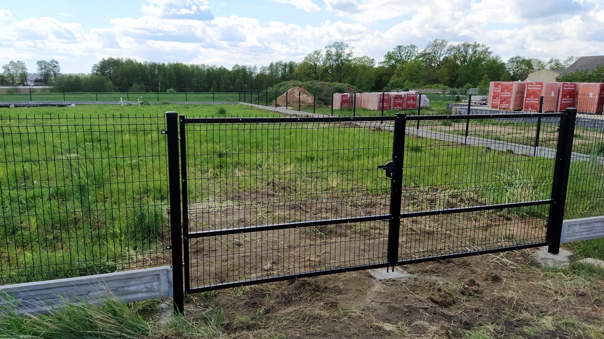 Czarna, metalowa furtka z siatki w ogrodzeniu panelowym, widok na trawnik i stosy czerwonej cegły w oddali.