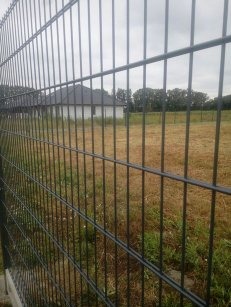 Ciemna siatka ogrodzeniowa z widocznym w tle białym domem i łąką pod pochmurnym niebem.