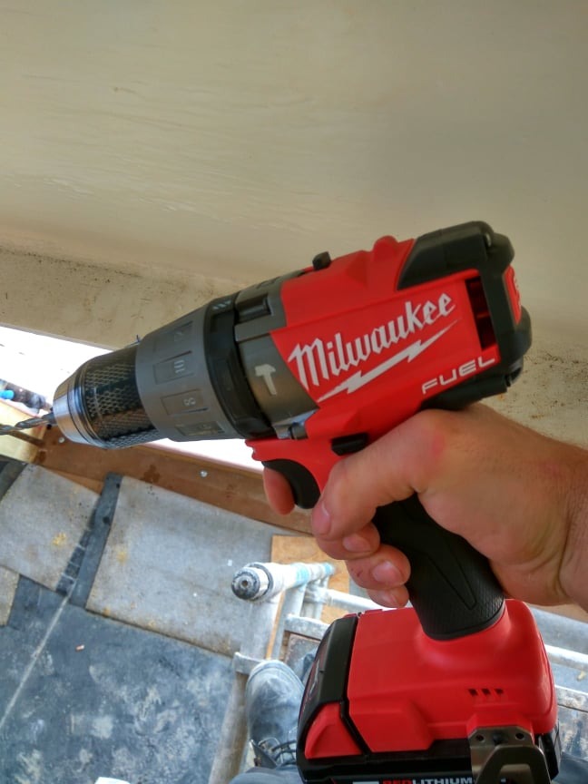 Czerwona wiertarko-wkrętarka Milwaukee Fuel w dłoni podczas wiercenia w suficie, widoczne fragmenty konstrukcji i but roboczy
