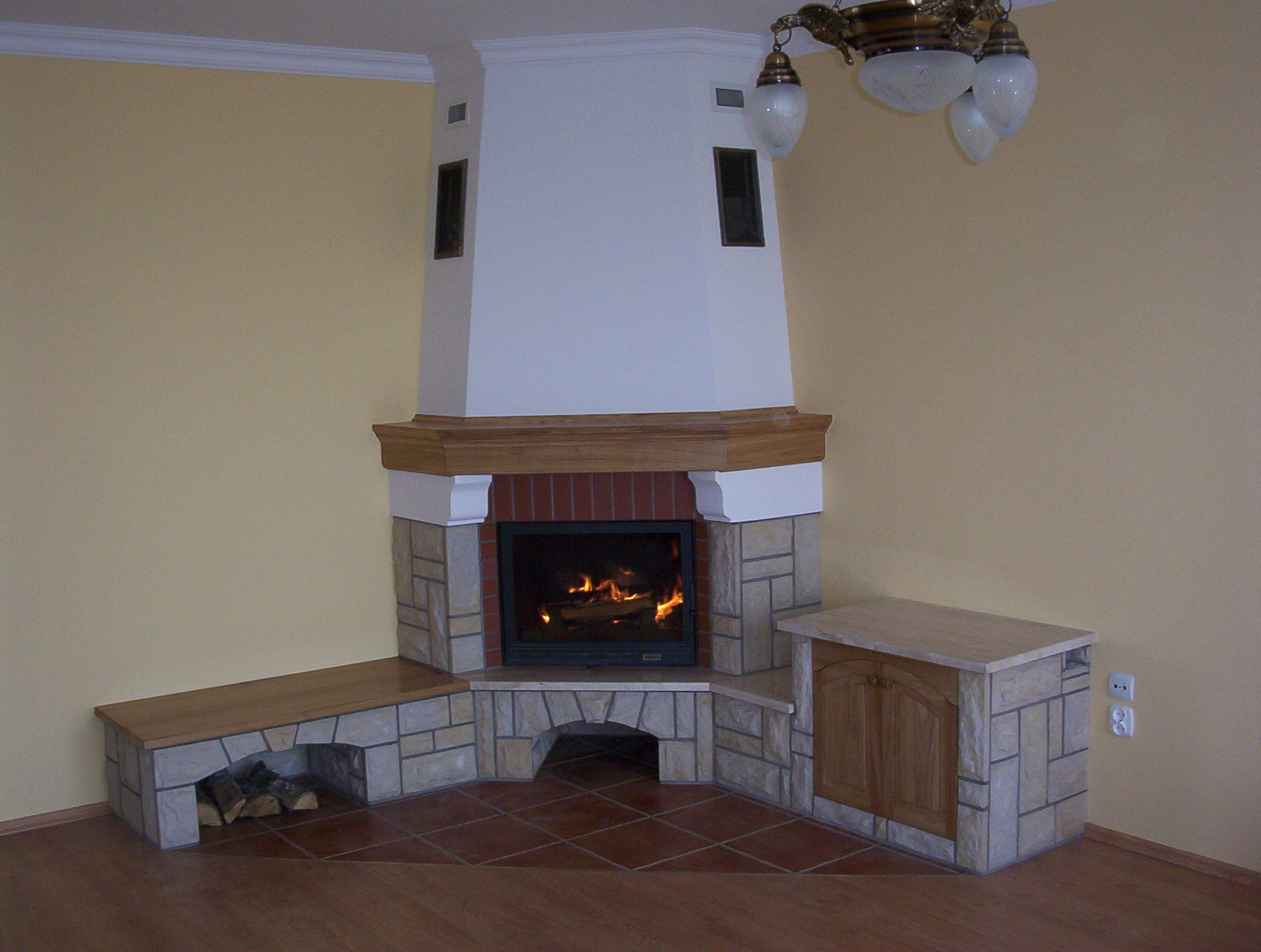 Kątowy wkład z paleniskiem obudowany kamieniem i drewnem, z drewnianą półką na drewno i szafką boczną, płomienie widoczne przez szybę.