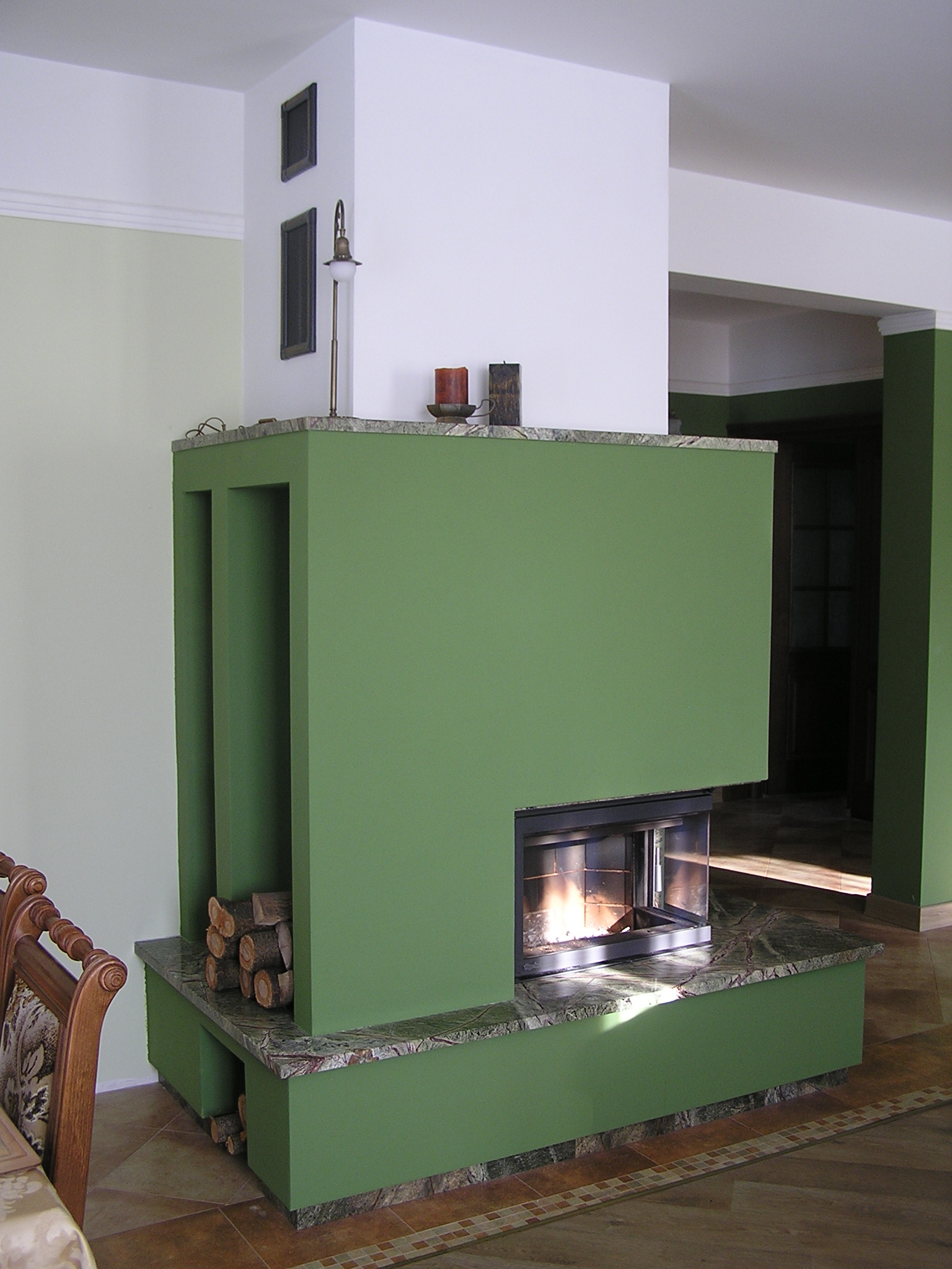 Nowoczesna zabudowa w odcieniach zieleni z marmurowym blatem, palenisko z widocznym płomieniem, drewno ułożone w niszach, wnętrze z elementami klasycznymi.