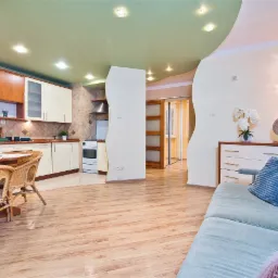 Jasne wnętrze mieszkania z drewnianą podłogą, otwartą kuchnią z beżowymi szafkami, okrągłym stołem z wiklinowymi krzesłami i turkusową sofą z poduszką.