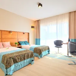 Jasny pokój hotelowy z dwoma łóżkami, drewnianym panelem na ścianie, stolikiem z krzesłami, zasłonami w odcieniach beżu i błękitu, oraz biurkiem z lampą