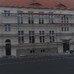Budynek w trakcie odświeżania elewacji, widoczne rusztowania, częściowo pomalowana fasada na biało, pracownicy i samoch&oacute;d dostawczy obok.