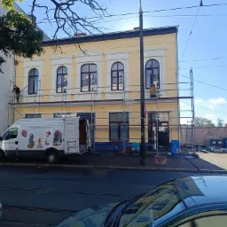 Dwupiętrowa, żółta kamienica z białymi zdobieniami wokół okien, w trakcie renowacji; dwóch pracowników na rusztowaniu, biały bus z grafikami narzędzi i prac na wysokości zaparkowany obok.