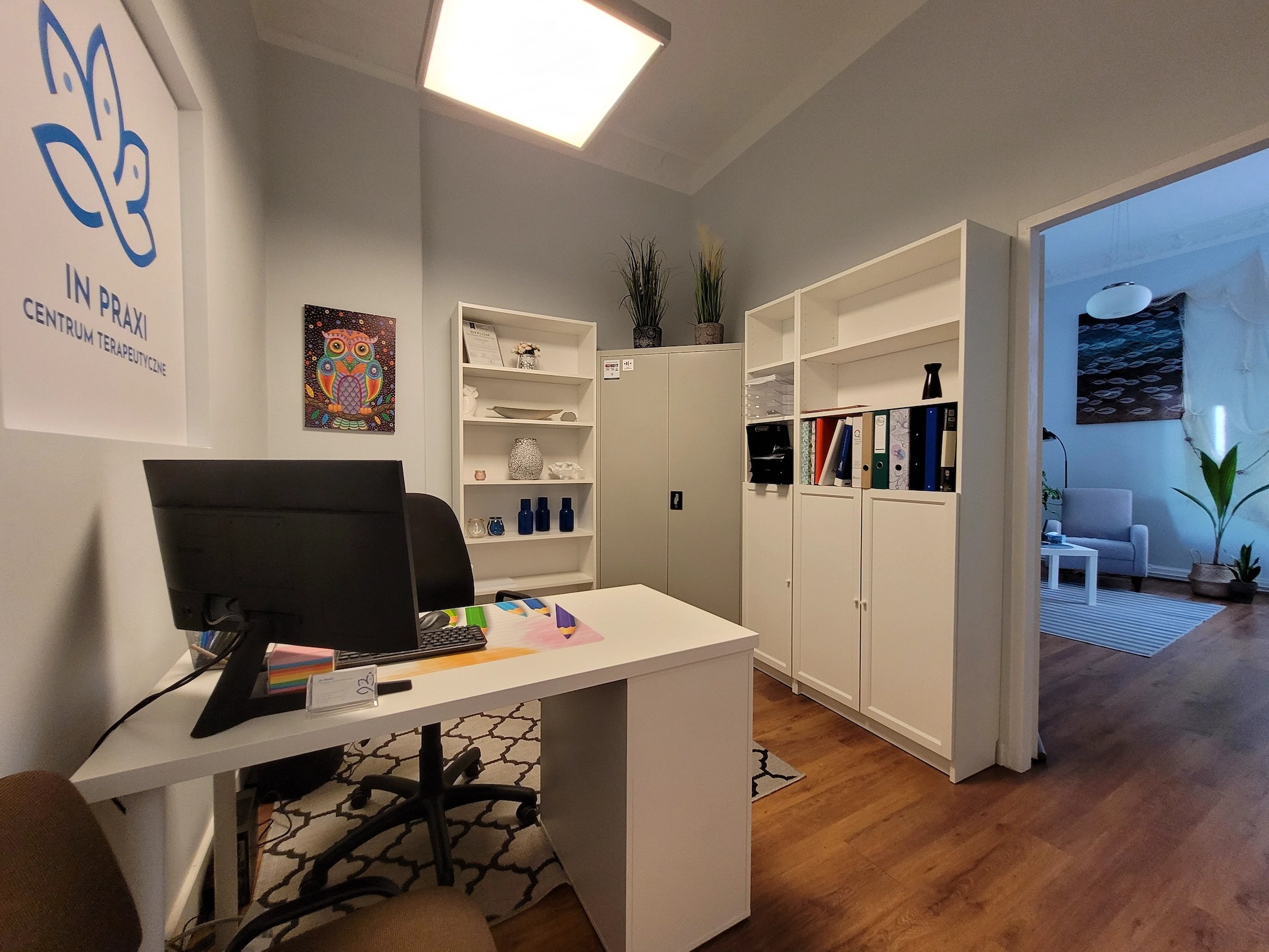 Jasny gabinet z biurkiem, komputerem i regałami, widoczne logo 'IN PRAXI Centrum Terapeutyczne' oraz pokój poczekalni w tle.
