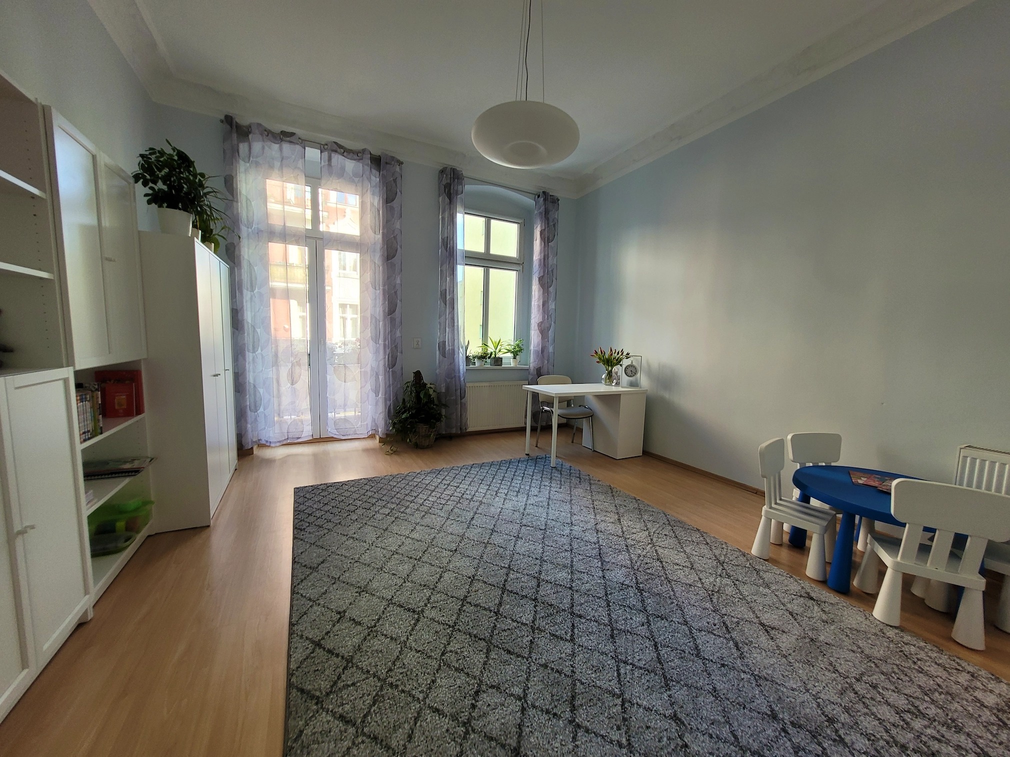 Jasny pokój z dywanem w romby, małym stolikiem i krzesłami dla dzieci, biurkiem pod oknem i regałem z książkami.