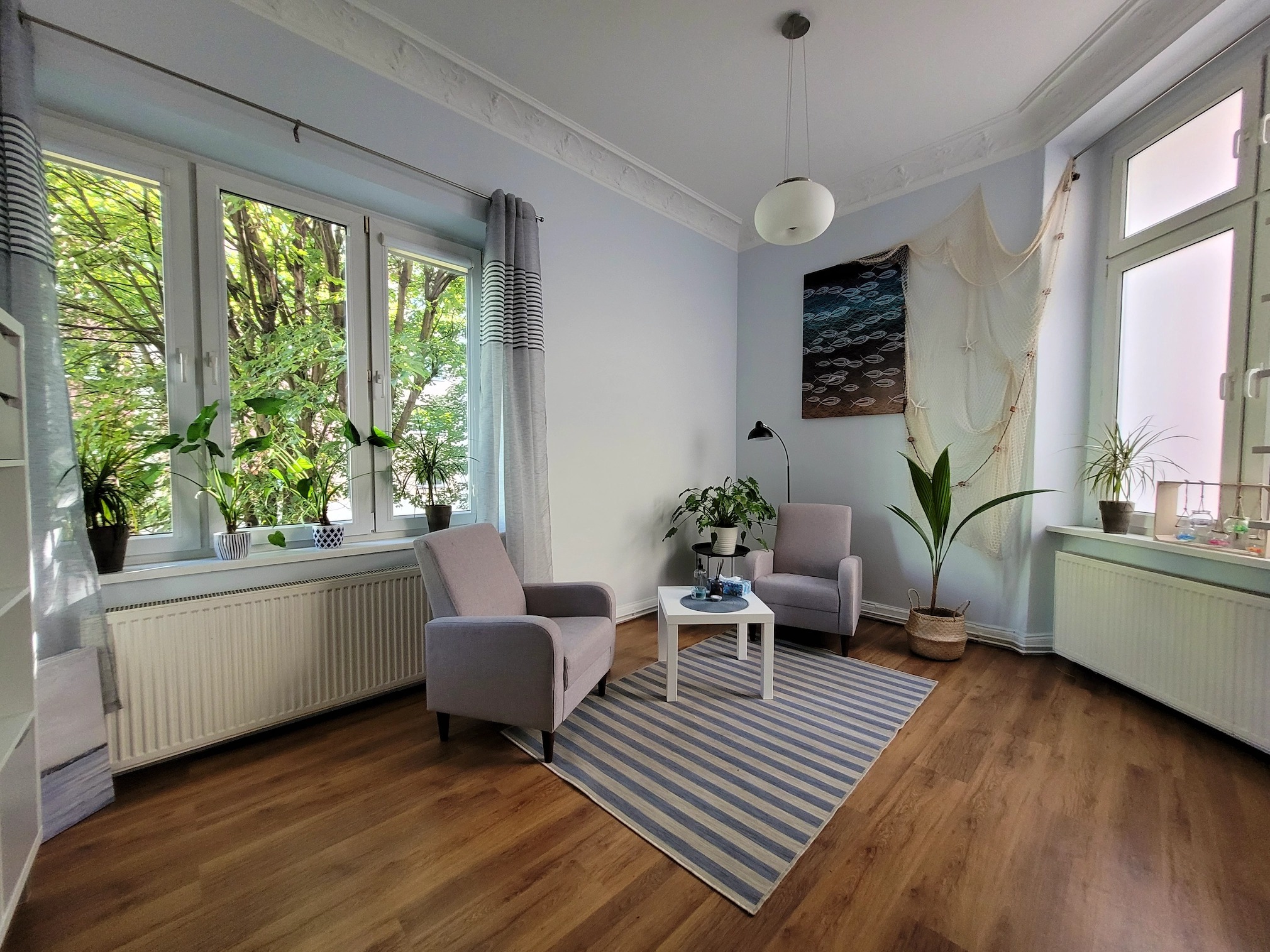 Jasny gabinet z dwoma fotelami, stolikiem kawowym, dywanem w paski, oknem z widokiem na zieleń i dekoracjami w stylu marynistycznym.