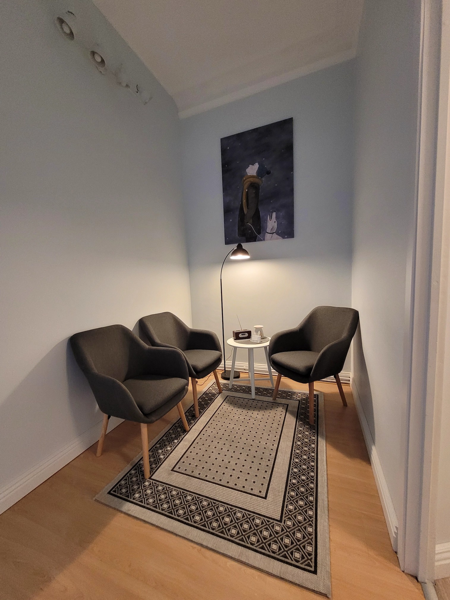 Gabinet terapeutyczny z trzema szarymi fotelami ustawionymi wokół małego białego stolika z lampką, dywanem w geometryczne wzory i obrazem na ścianie, tworzący spokojną atmosferę.
