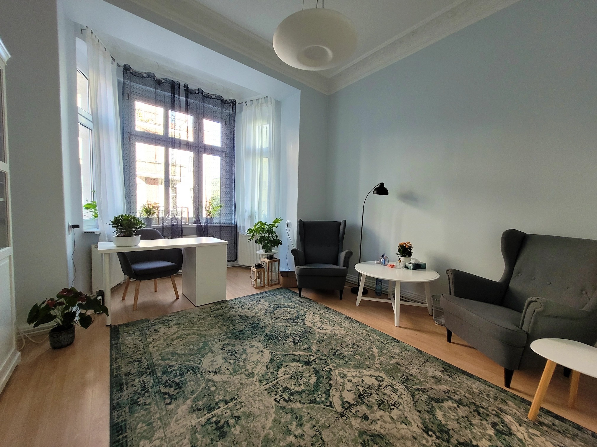 Jasny gabinet z dużym oknem, dwoma fotelami typu uszak, białym biurkiem i dywanem w odcieniach zieleni i beżu, lampa podłogowa i sufitowa