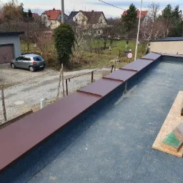 Widok z góry na płaski dach z brązowymi, metalowymi elementami wykończeniowymi, na tle zabudowań mieszkalnych i garażu, z deskami i drewnianymi klockami leżącymi na powierzchni dachu.