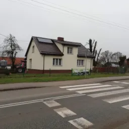 Dwukondygnacyjny dom jednorodzinny z ciemnobrązowym dachem i panelami słonecznymi, widziany z ulicy z przejściem dla pieszych, traktor w tle.