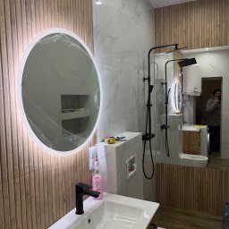 KRISPOL Krystian Żuk - Nowoczesna łazienka po remoncie: okrągłe lustro LED w folii ochronnej, drewnopodobne płytki ścienne, biała umywalka z czarną armaturą i widoczne elementy instalacji.