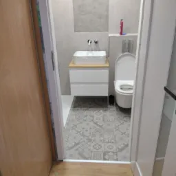 Nowoczesna łazienka po remoncie: umywalka nablatowa na szafce z szufladami, lustro wnękowe, toaleta i wzorzyste płytki podłogowe widziane zza uchylonych drzwi.