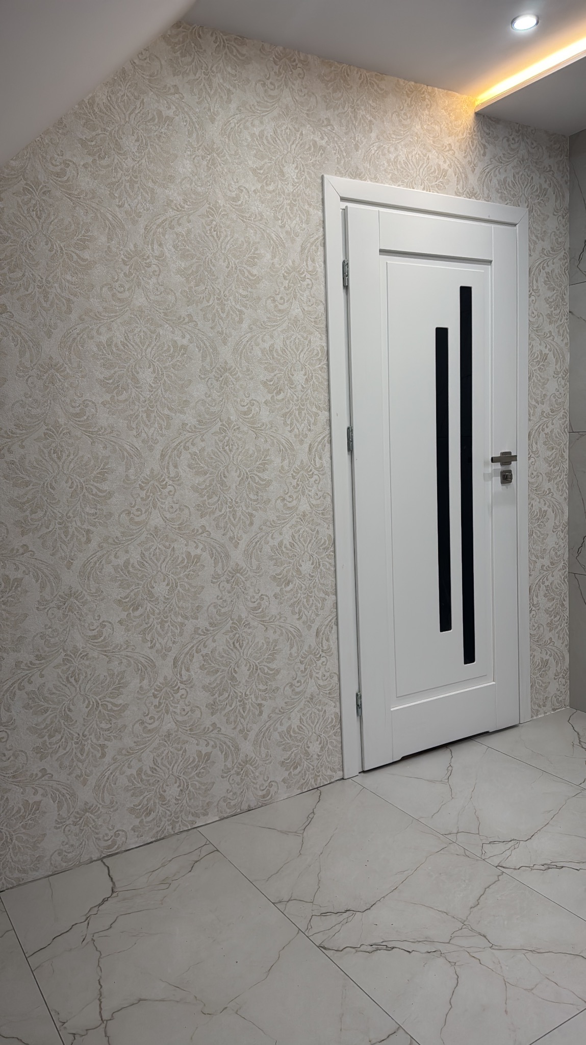Tapeta w kremowe ornamenty na ścianie, białe drzwi z czarnymi wstawkami i marmurowe płytki podłogowe. Nowoczesne wykończenie wnętrza.