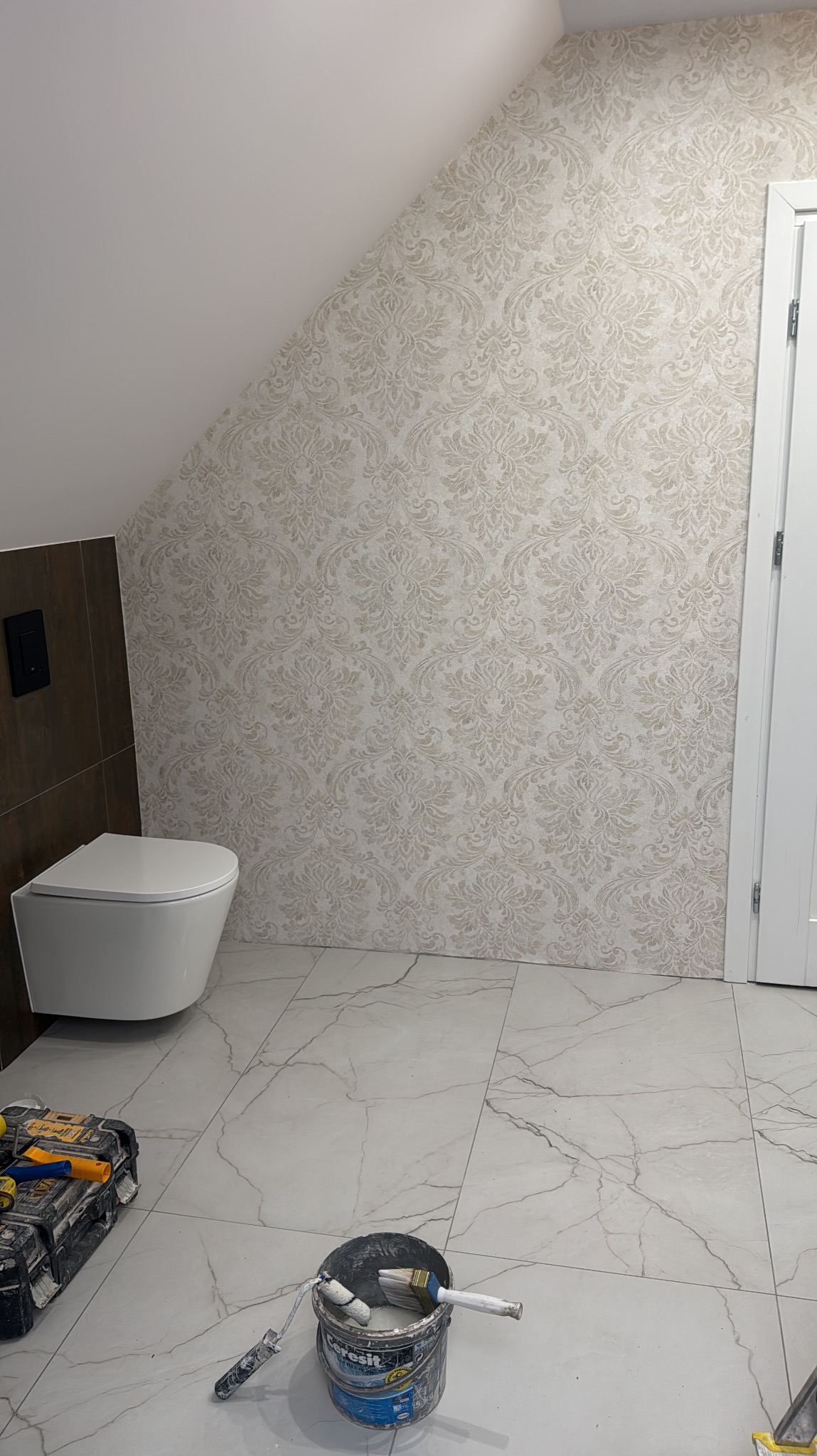 Ukończona łazienka na poddaszu z wzorzystą tapetą w odcieniach beżu, białą ceramiką i marmurową podłogą. Widoczne narzędzia malarskie.