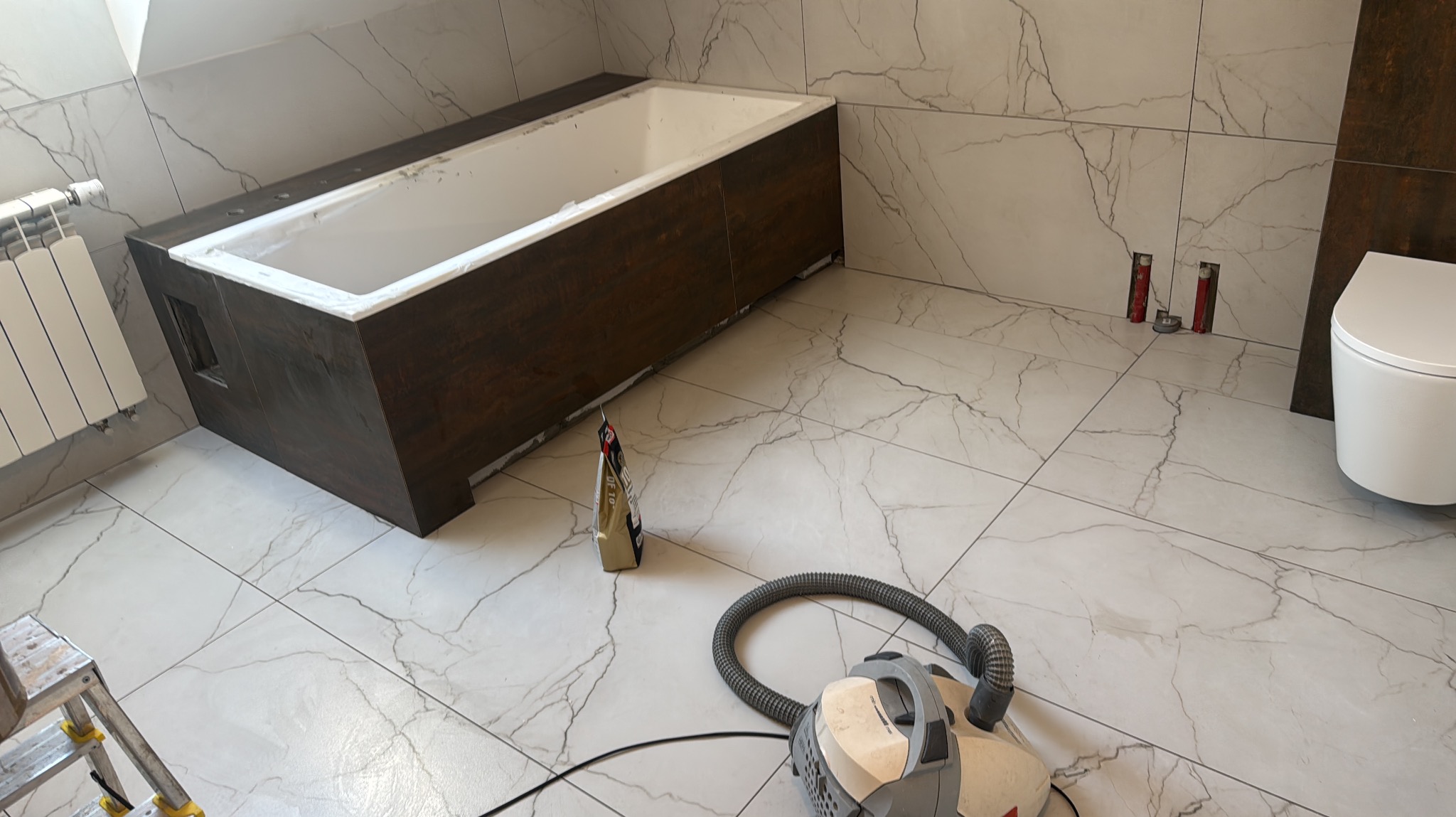 Nowoczesna łazienka w trakcie wykańczania: białe płytki imitujące marmur na podłodze i ścianach, obudowana wanna z ciemnego drewna, biała toaleta i odkurzacz.