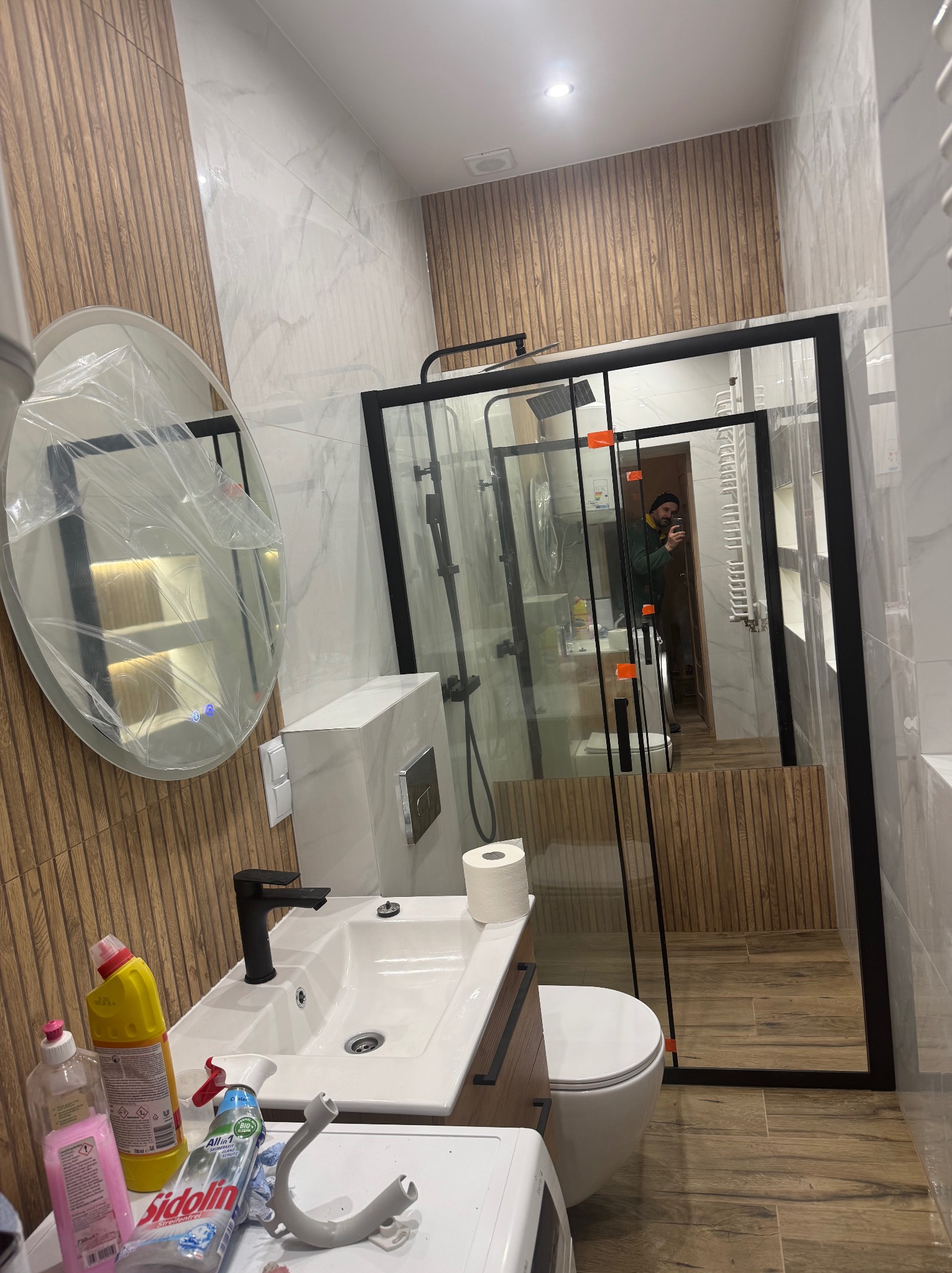 Nowoczesna łazienka po remoncie: drewnopodobne płytki, kabina prysznicowa z czarną ramą, umywalka z szafką i okrągłe lustro z podświetleniem, częściowo zakryte folią ochronną.