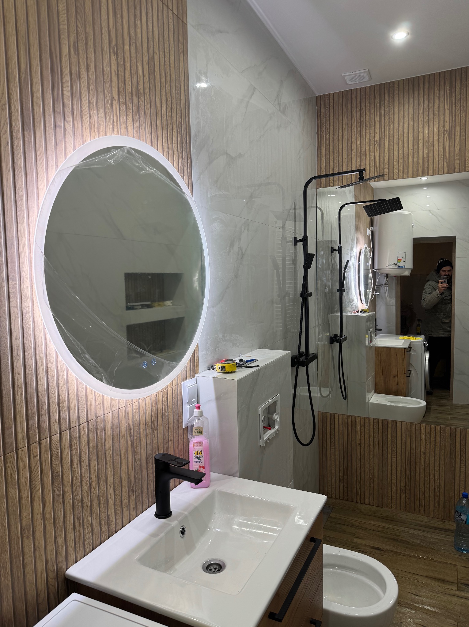 Nowoczesna łazienka po remoncie: okrągłe lustro LED w folii ochronnej, drewnopodobne płytki ścienne, biała umywalka z czarną armaturą i widoczne elementy instalacji.