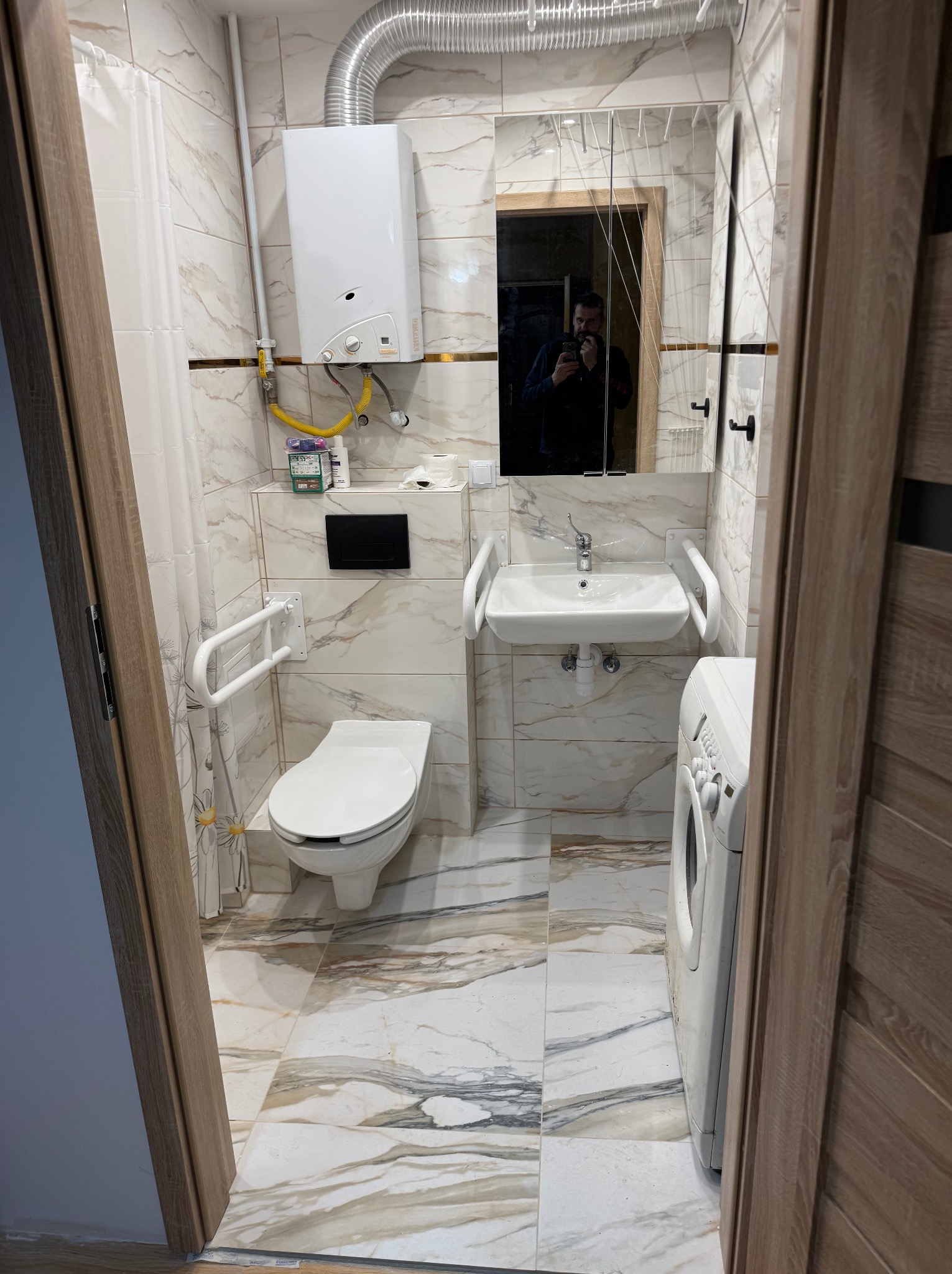 Wykończona łazienka z marmurowymi płytkami, umywalką z uchwytami dla niepełnosprawnych, toaletą, pralką i lustrem z odbiciem osoby robiącej zdjęcie.