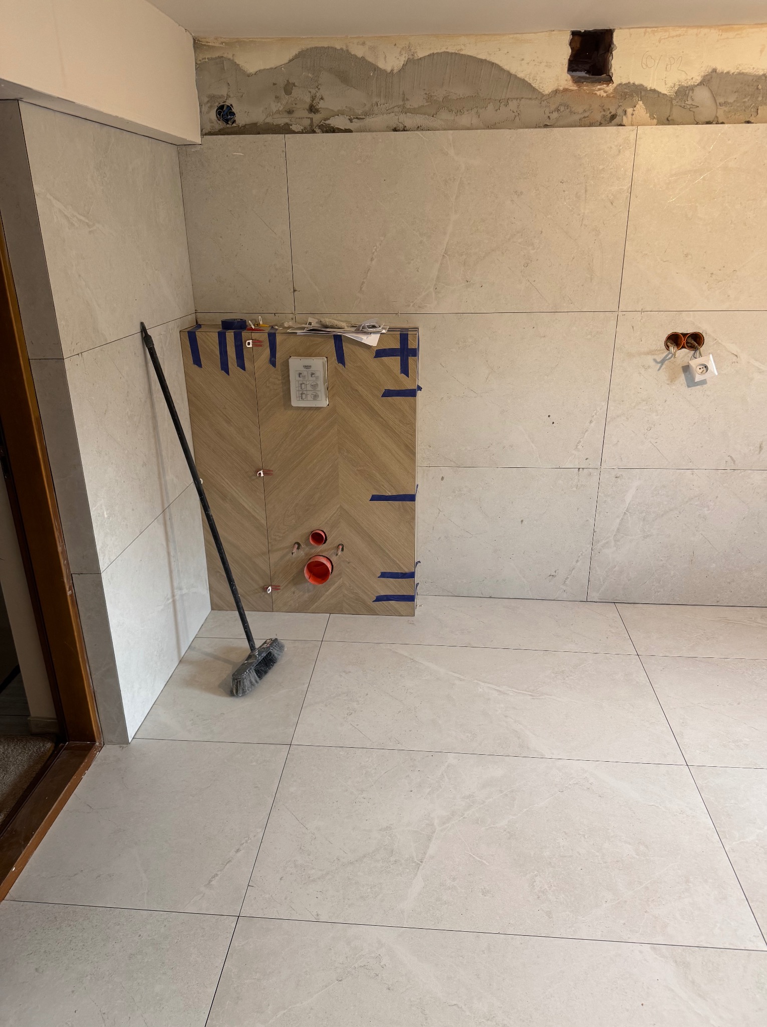 Jasne płytki w łazience w trakcie montażu, widoczne fragmenty instalacji i przygotowane miejsce pod zabudowę. Na podłodze leży zmiotka.