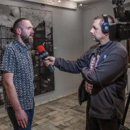 Wywiad z mężczyzną na tle czarno-białej fototapety, mikrofon z logo TVK w kadrze, kamera na statywie po prawej stronie.