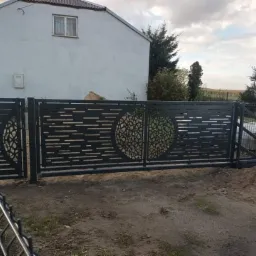 Nowoczesna, przesuwna brama wjazdowa z metalu, z ażurowym wzorem geometrycznym w kształcie koła, na tle domu jednorodzinnego i słupa energetycznego.