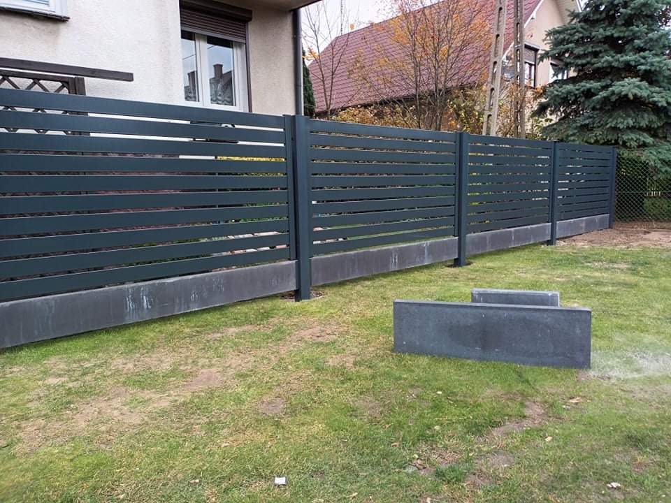 Nowoczesne ogrodzenie z poziomych, ciemnoszarych paneli metalowych na betonowej podmurówce, widoczne elementy montażowe i przygotowane bloczki betonowe na trawniku.