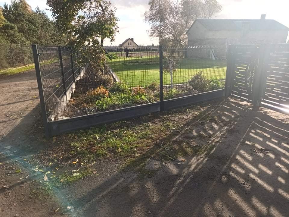 Zamontowane ogrodzenie panelowe z bramą wjazdową na posesji z widocznym domem w tle, ziemia wokół ogrodzenia z liśćmi i śladami opon.