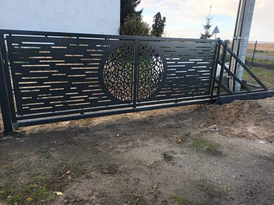 Nowoczesna, przesuwna brama metalowa w kolorze antracytowym z dekoracyjnym, ażurowym wzorem w kształcie koła i poziomych pasków, widoczna na tle pola i słupa energetycznego.