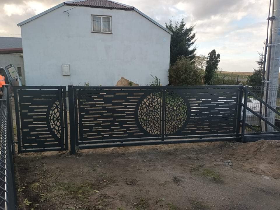 Nowoczesna, metalowa brama wjazdowa z ażurowym wzorem koła na tle białego domu jednorodzinnego i pochmurnego nieba.