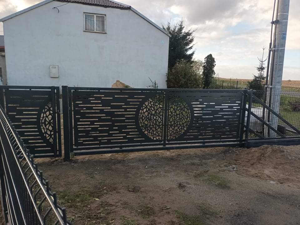 Nowoczesna, przesuwna brama wjazdowa z metalu, z ażurowym wzorem geometrycznym w kształcie koła, na tle domu jednorodzinnego i słupa energetycznego.