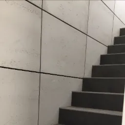 Klatka schodowa z szarymi schodami i ścianą wyłożoną jasnymi płytami imitującymi trawertyn, oddzielonymi czarnymi listwami.