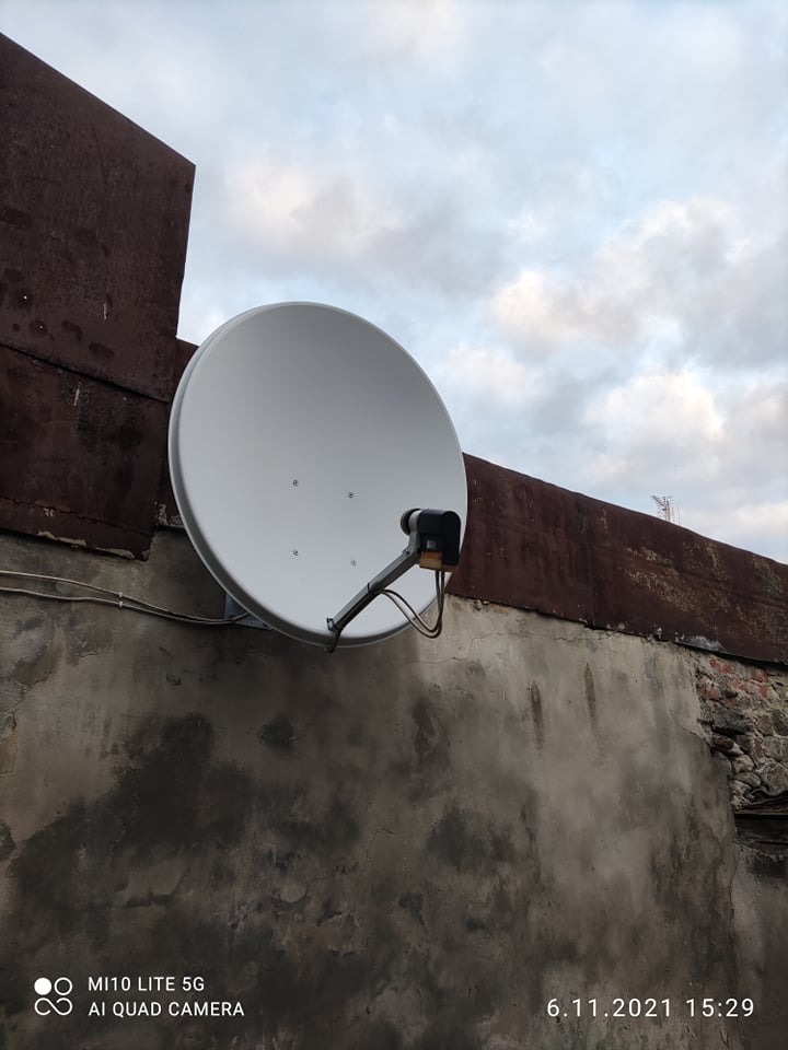 Biała antena satelitarna zamontowana na szarej, popękanej ścianie budynku z fragmentami cegieł, na tle pochmurnego nieba.