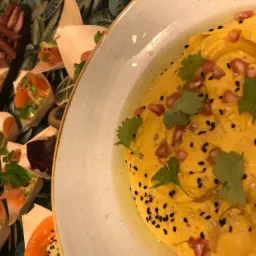 Żółta pasta z posypką z czarnego i białego sezamu oraz granatu, udekorowana zieloną pietruszką, obok przekąski w drewnianych łódeczkach na tle wzorzystej serwety.