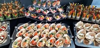 Elegancko zaaranżowany stół z różnorodnymi przekąskami: mini sałatki w szklanych naczyniach na wielopoziomowym stojaku, finger food w papierowych łódeczkach z łososiem, warzywami i oliwkami,...