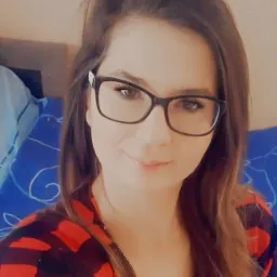 Portret kobiety w okularach z brązowymi włosami, uśmiechniętej, ubranej w czerwoną koszulę w czarną kratę, na tle niebieskiej pościeli.