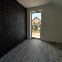 Pokój na poddaszu z szarą podłogą z paneli, czarną ścianą z geometrycznym wzorem i widokiem na dom przez okno, widoczna listwa przypodłogowa.