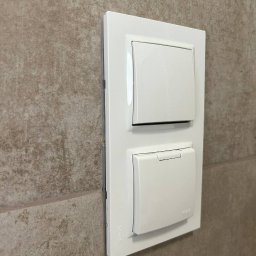 OPTIMA SKY - Biały podwójny włącznik światła Simon na beżowej, teksturowanej ścianie. Dolny włącznik z klapką. Ujęcie z boku, widoczny cień. Minimalistyczny design.