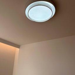 OPTIMA SKY - Nowoczesna, okrągła lampa sufitowa LED z białym kloszem, zamontowana na jasnym suficie w minimalistycznym wnętrzu. Widoczne gniazdka elektryczne na ścianie.