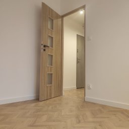 OPTIMA SKY - Otwarta, drewniana drzwi wewnętrzne z przeszkleniami, widoczne na tle parkietu ułożonego w jodełkę i białych ścian z listwami przypodłogowymi. Widok na kolejne pomieszczenie.