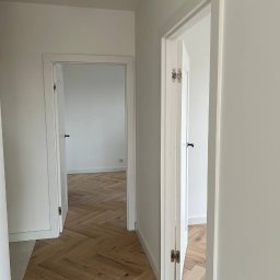 OPTIMA SKY - Jasne wnętrze z parkietem w jodełkę, widok na otwarte, białe drzwi. Minimalistyczny design, neutralna kolorystyka, subtelne oświetlenie punktowe.