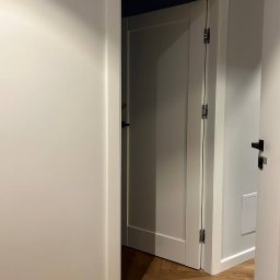 OPTIMA SKY - Otwarta, biała, wewnętrzna stolarka drzwiowa z widocznymi zawiasami, na tle jasnych ścian i parkietu ułożonego w jodełkę. Minimalistyczny design.