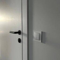 OPTIMA SKY - Białe drzwi z czarną klamką i włącznik światła na ścianie. Minimalistyczny design, neutralna kolorystyka, nowoczesne wykończenie. Detal wnętrza po remoncie.