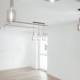 OPTIMA SKY - Jasne wnętrze po remoncie: panele podłogowe, białe ściany z widocznymi gniazdkami, nowoczesne lampy wiszące. Minimalistyczny design, naturalne światło.