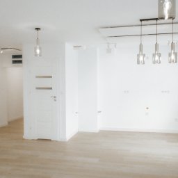 OPTIMA SKY - Jasne, przestronne wnętrze po remoncie: parkiet, białe ściany, nowoczesne lampy wiszące. Widoczne gniazdka elektryczne i drzwi wewnętrzne. Minimalistyczny design.