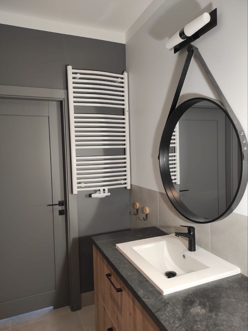 Nowoczesna łazienka w szarościach: biały grzejnik drabinkowy, okrągłe lustro z czarną ramą, umywalka z czarną armaturą i drewniana szafka pod umywalką.