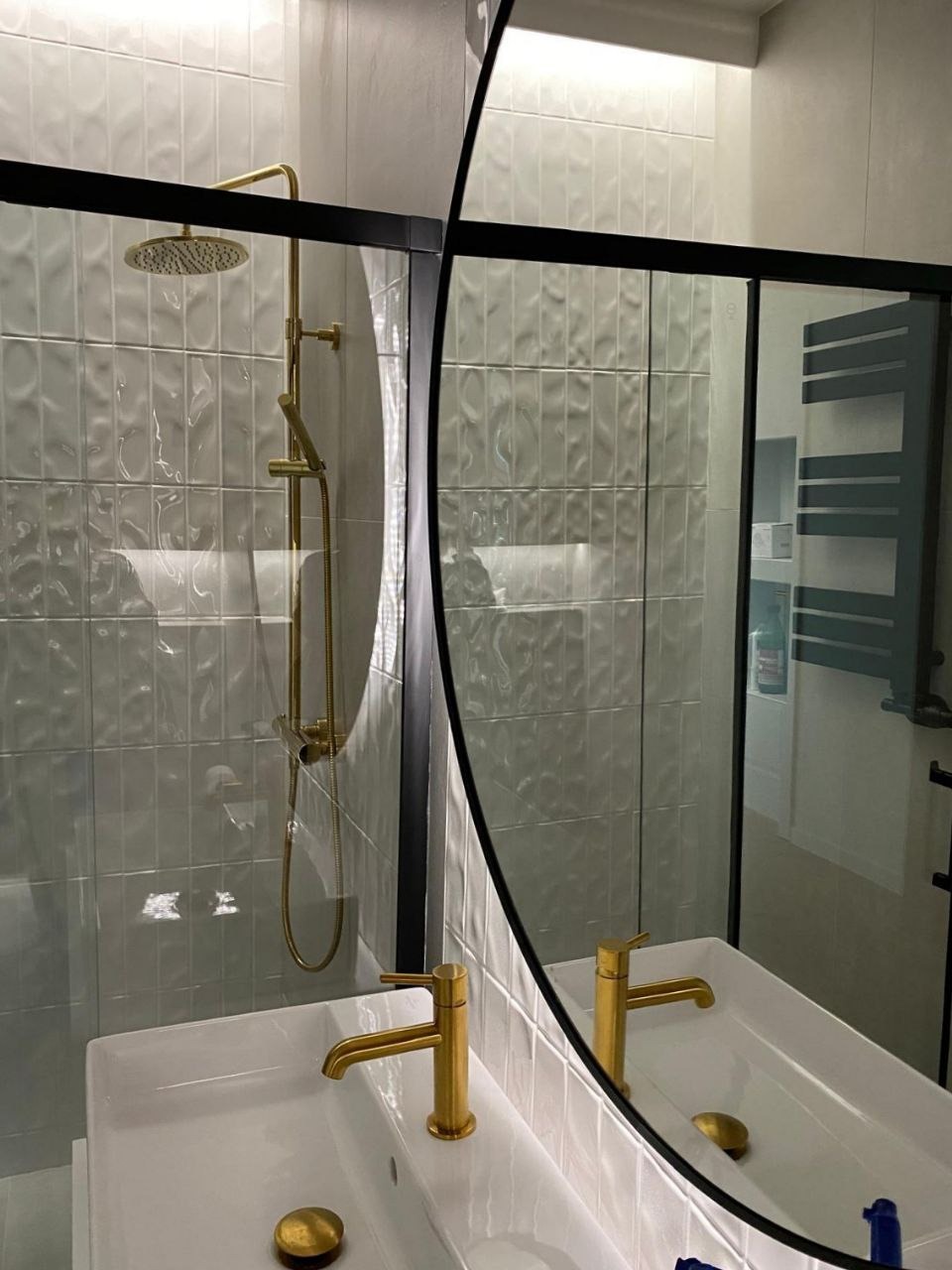 Nowoczesna łazienka z kabiną prysznicową typu walk-in, białą umywalką i złotą armaturą, odbijająca się w okrągłym lustrze z czarną ramą.